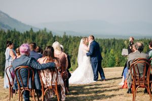 Bride and groom share first kiss at outdoor mountain ceremony with forest and rolling hills backdrop.