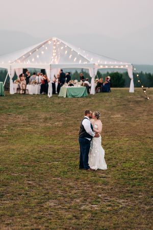 Bride and groom share a romantic kiss in a green field with a beautifully lit wedding reception tent in the background.