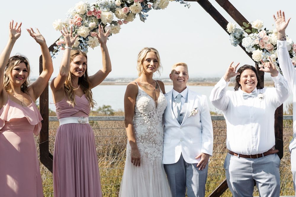Joyful wedding party celebrating outdoors near water under floral arch, bride in white gown with bridesmaids and guests.