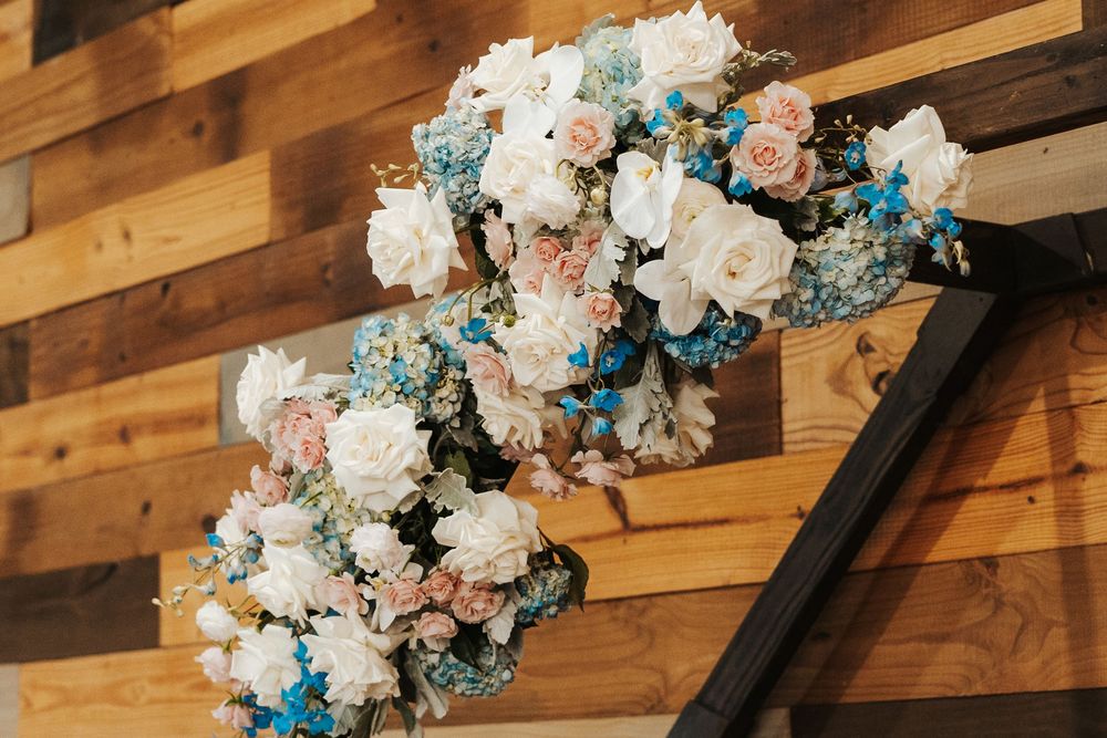 Elegant wedding arch adorned with white and pink roses, blue flowers, and baby's breath against a rustic wood wall.