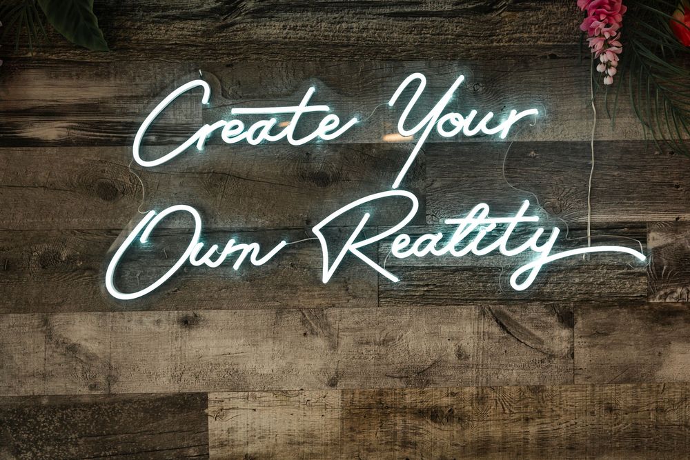 Neon sign reading Create Your Own Reality mounted on a rustic wooden wall with floral decor.