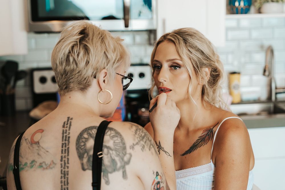 Tattoo artist applying red lipstick to a bride during wedding makeup preparation in a modern kitchen.