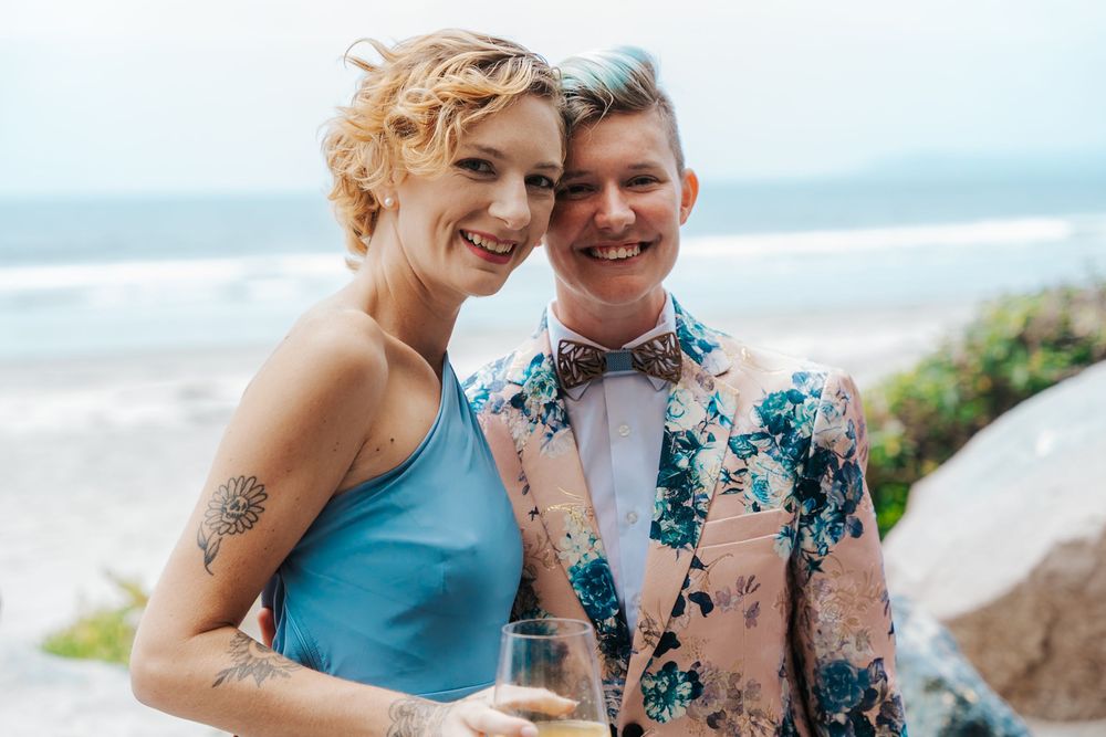 Smiling couple at beach wedding, woman in blue dress with tattoos, man in floral suit with bow tie.