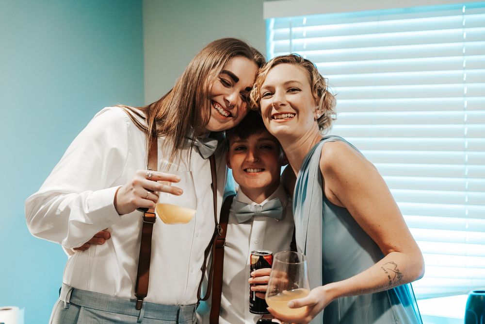 Three friends smiling and holding drinks together at a celebration, dressed in formal attire.