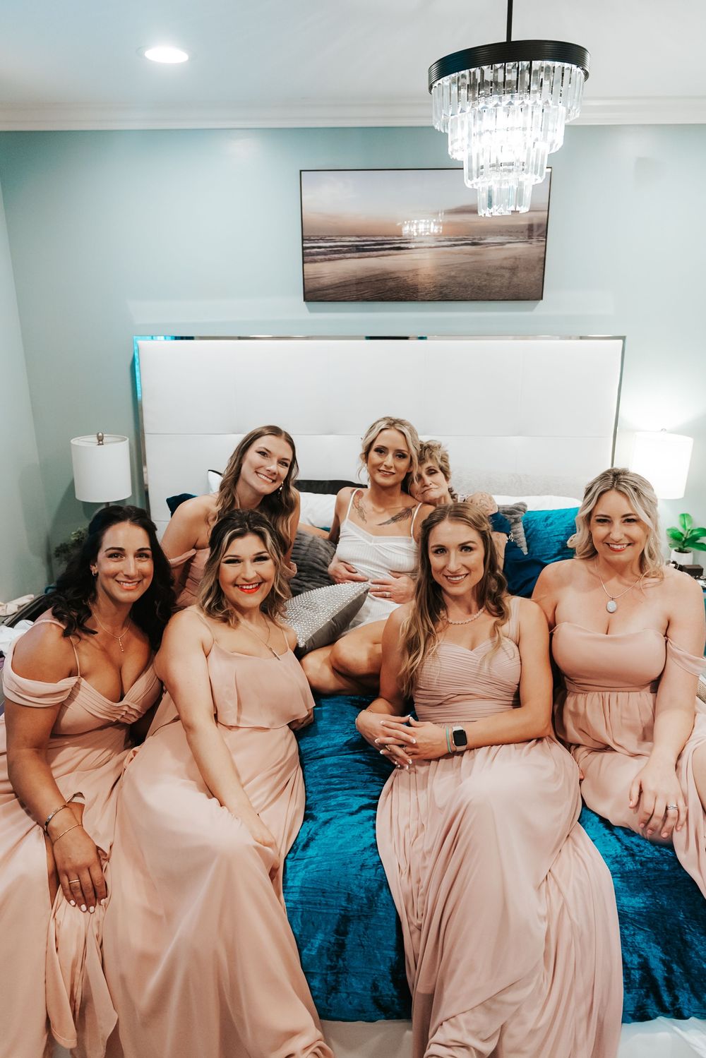 Six bridesmaids in blush pink gowns pose on a teal bed with a bride in a white dress in an elegant bedroom.