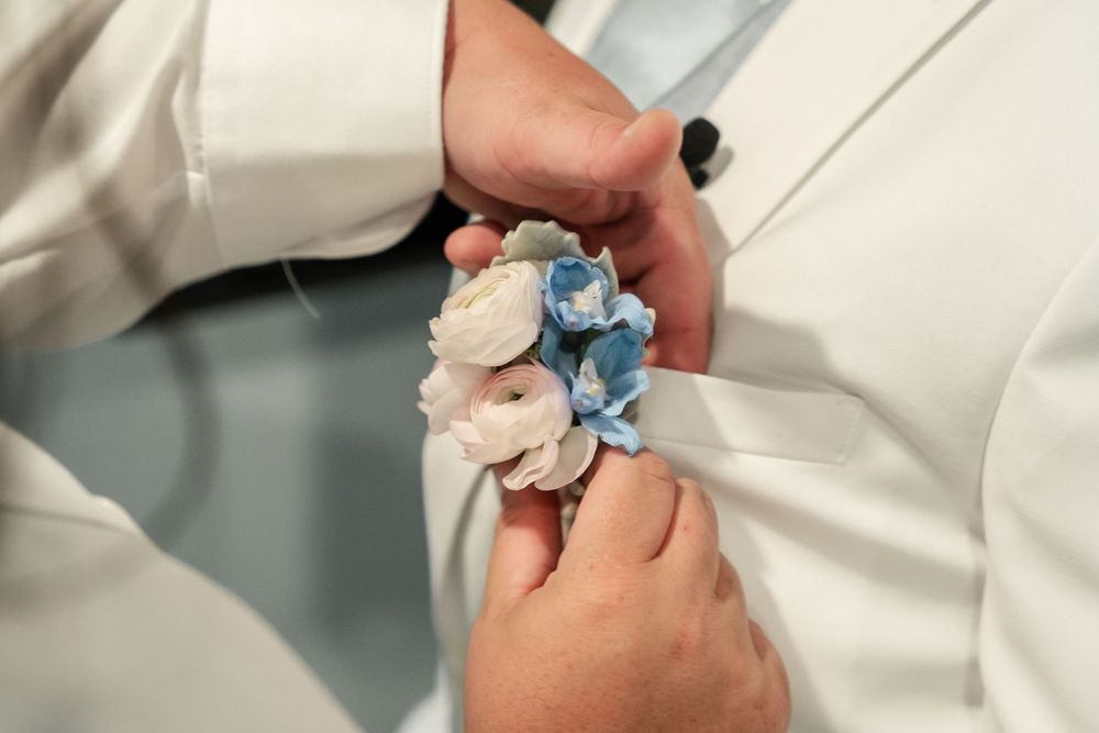 Close-up of hands pinning blue and white flower boutonniere on white suit lapel at wedding ceremony.