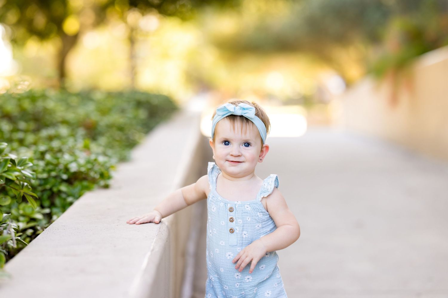 Adorable toddler girl in blue romper and bow headband standing on outdoor pathway surrounded by greenery.