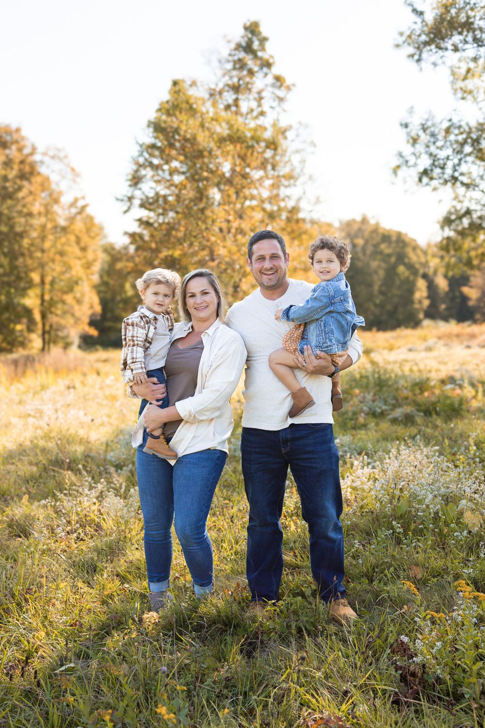 Family of four posing outdoors in a sunny autumn field, parents holding two young children, wearing casual jeans and light tops.