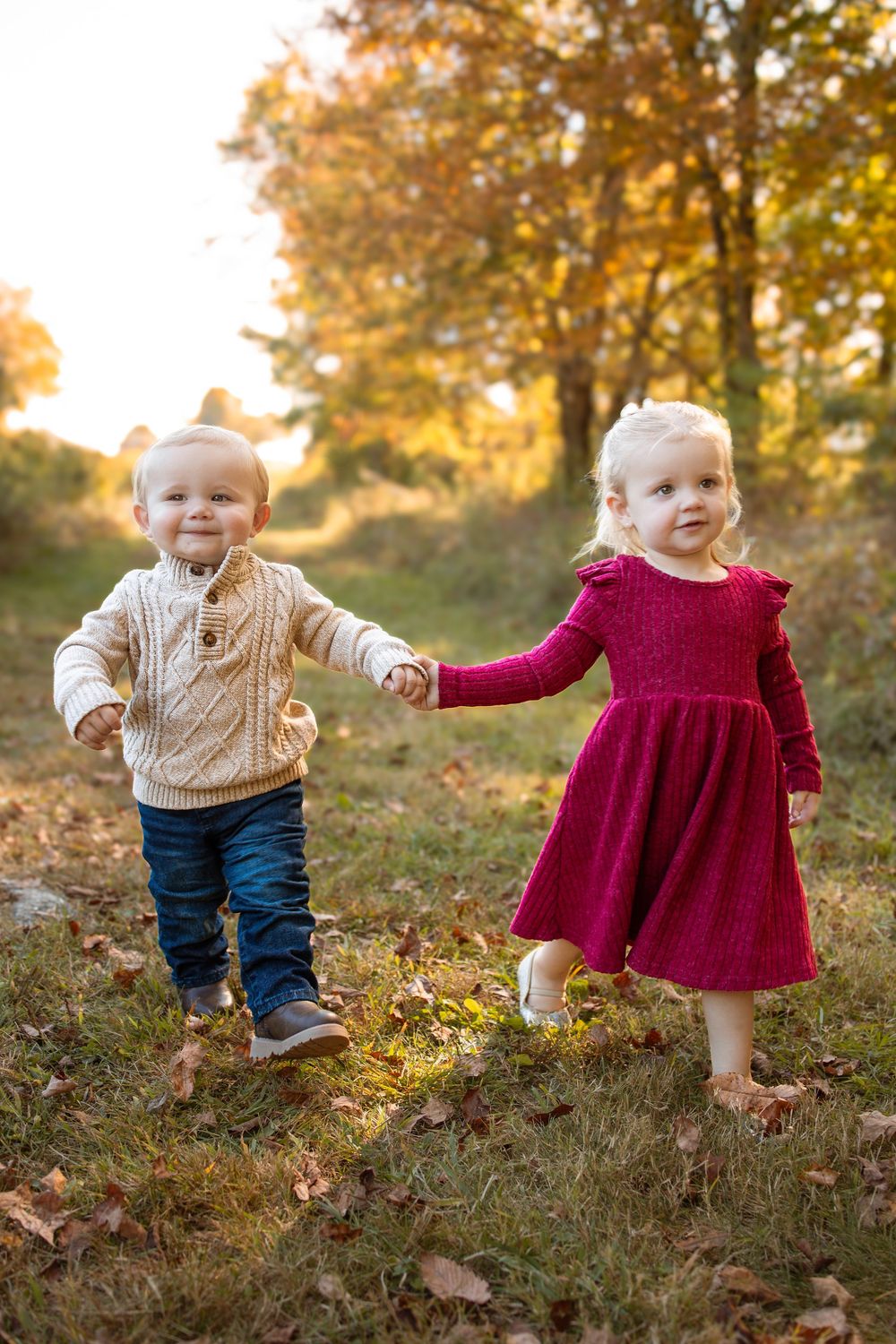 Two toddlers holding hands in autumn park, boy in cream sweater and jeans, girl in red dress, golden fall foliage background.