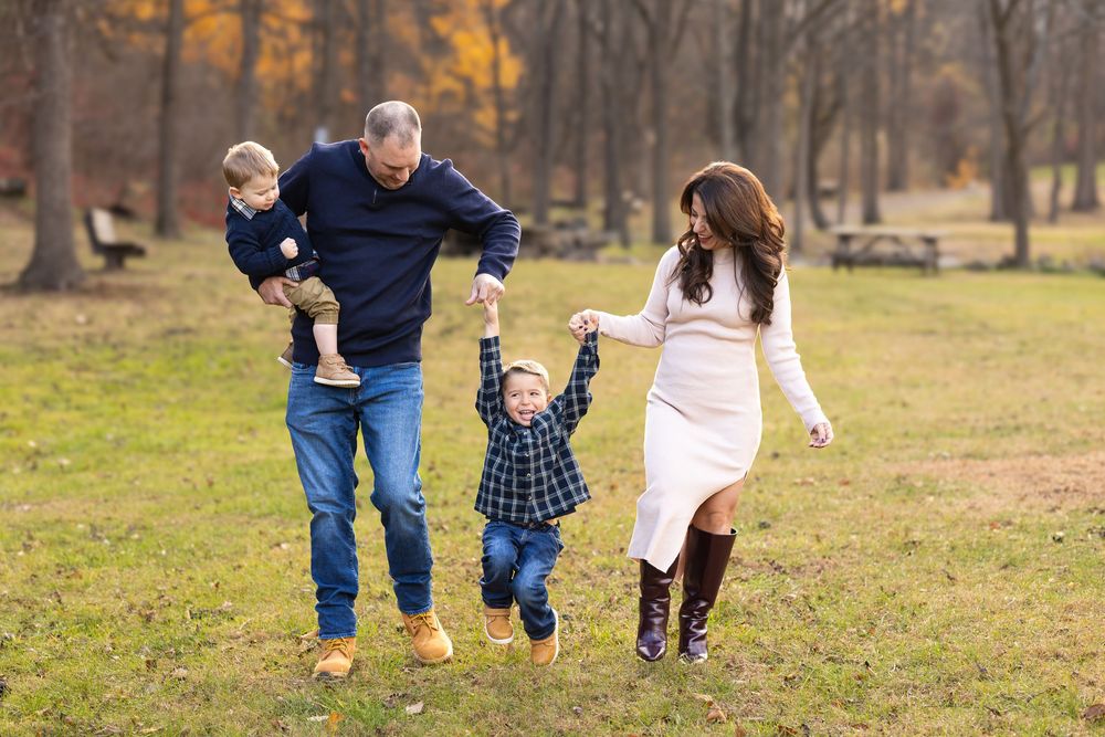 Family of four walking in autumn park, parents swinging young boy between them, toddler on dad's hip, fall foliage background.