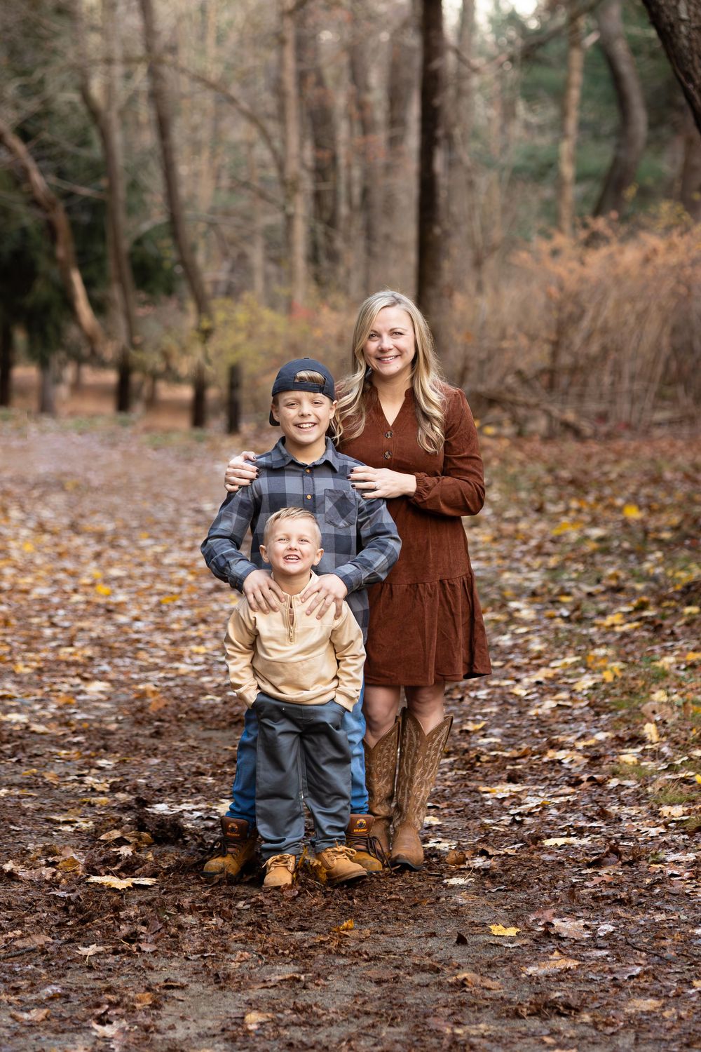 A woman in a brown dress and boots poses with two young boys on a leaf-covered forest path in autumn.
