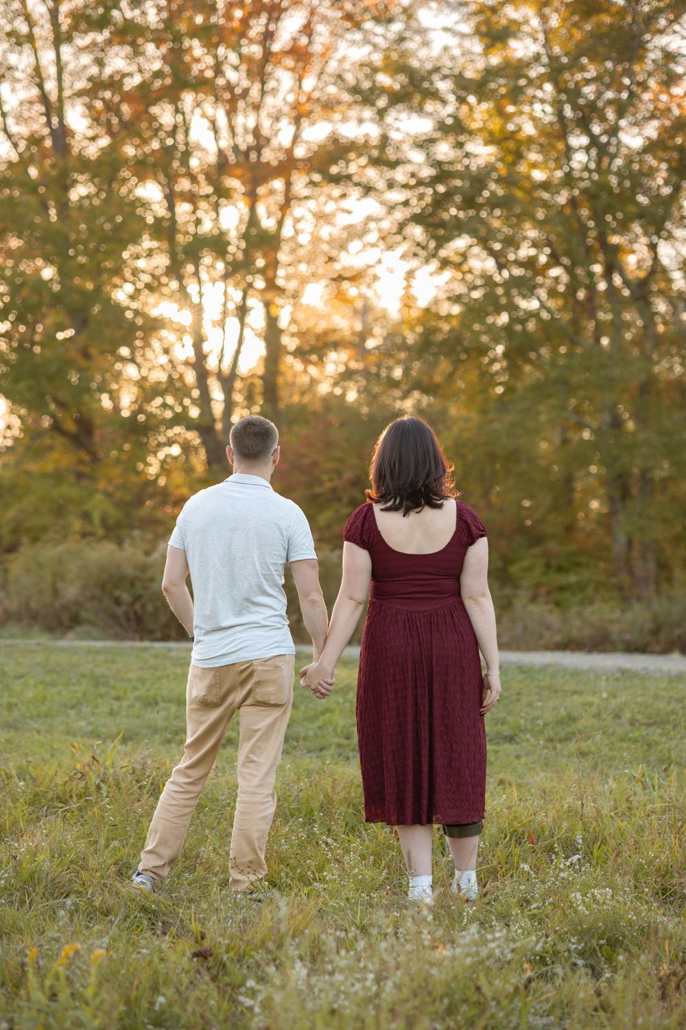 Couple holding hands walking through a grassy field at golden hour with autumn trees in background.