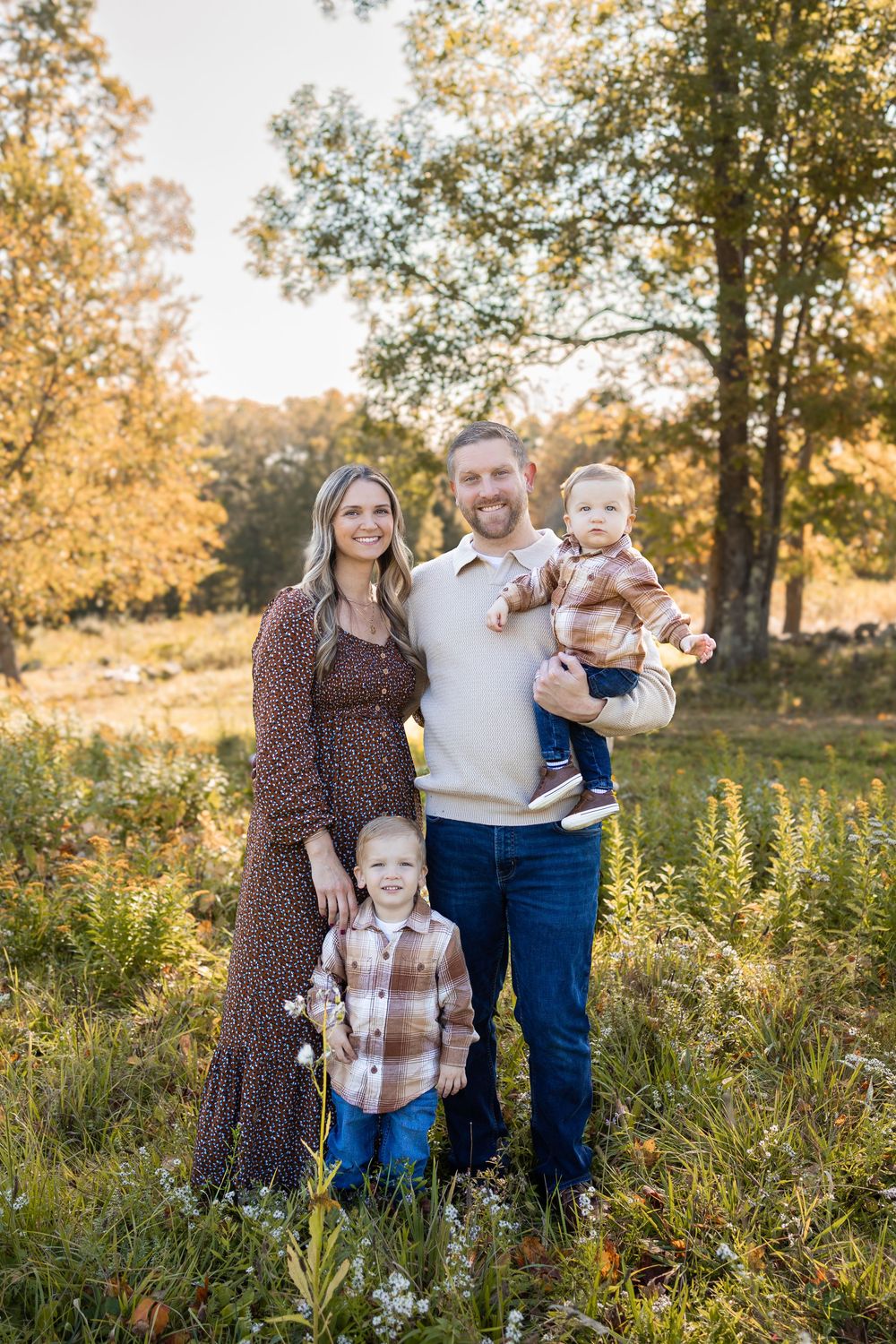 Family of four posing outdoors in autumn, parents with two young children smiling in a sunlit meadow with fall foliage.
