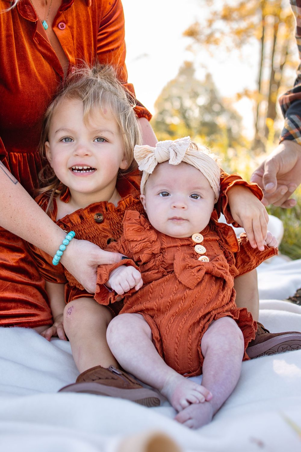 Two young sisters in matching rust-colored outfits sit together outdoors, the older girl smiling brightly beside her baby sister.