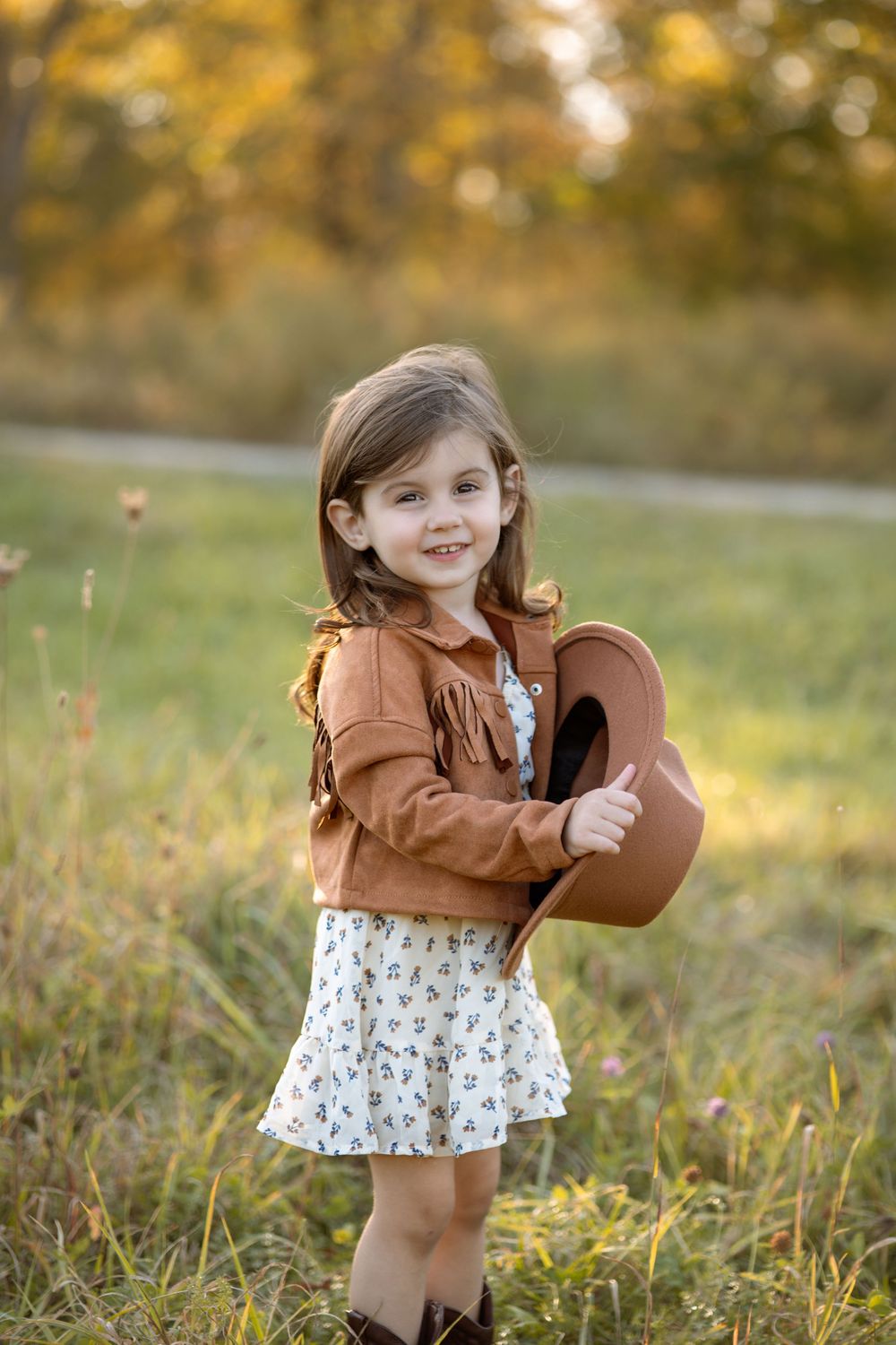 A smiling little girl in a brown fringe jacket, floral dress, and cowboy boots holds a hat in a sunny field.