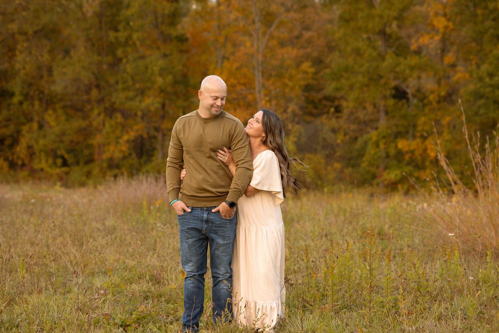Couple standing in autumn field, man in green sweater and jeans, woman in cream dress, surrounded by fall foliage.