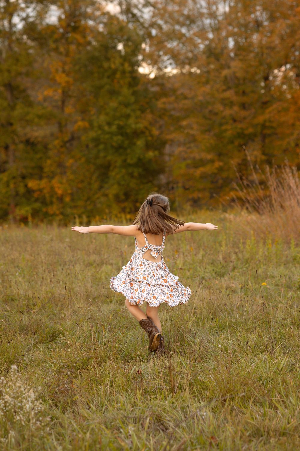 A young girl in a floral dress and boots twirls joyfully in an autumn meadow with colorful trees in the background.