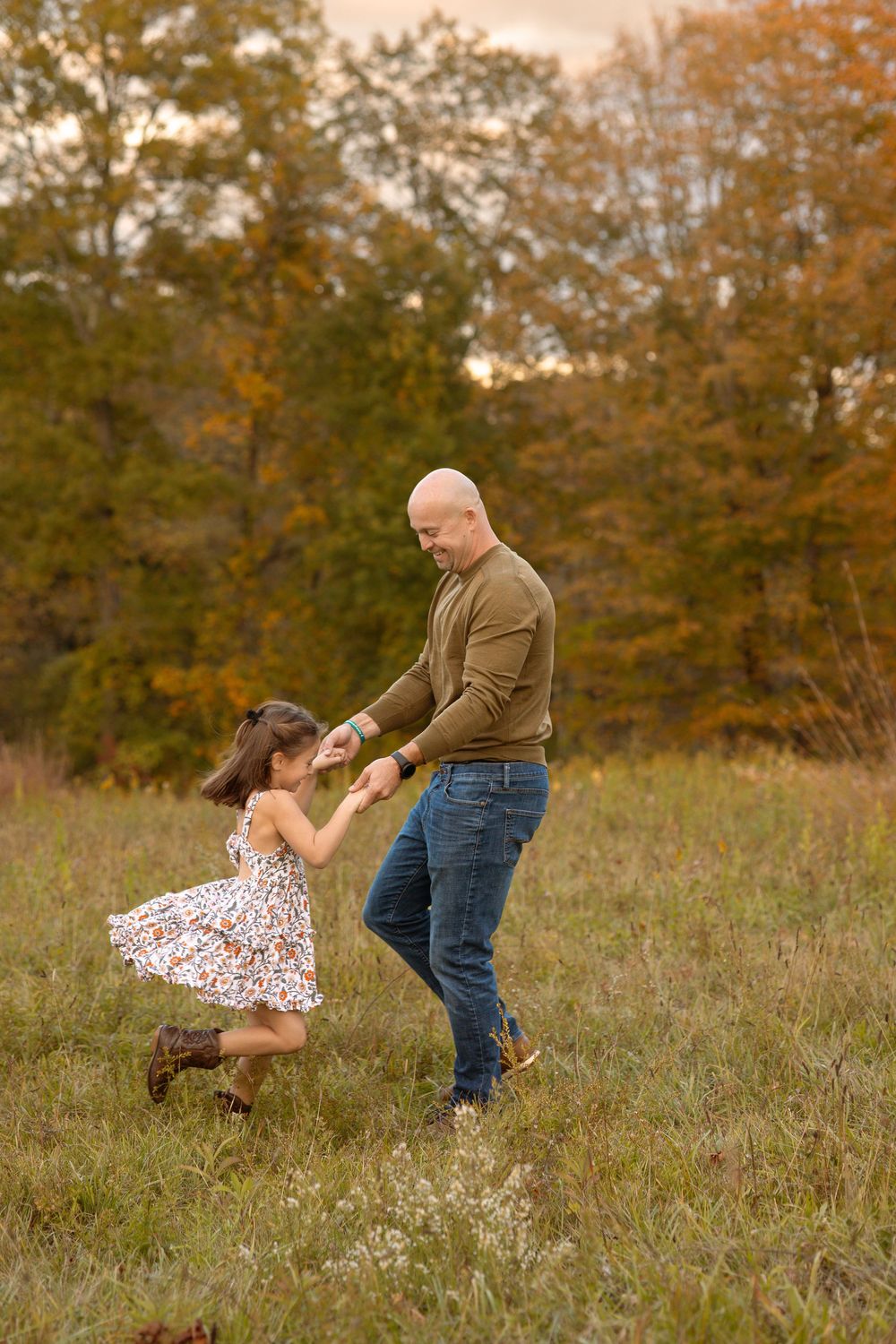 A bald father dances with his young daughter in a field of autumn foliage during a fall family photo session.
