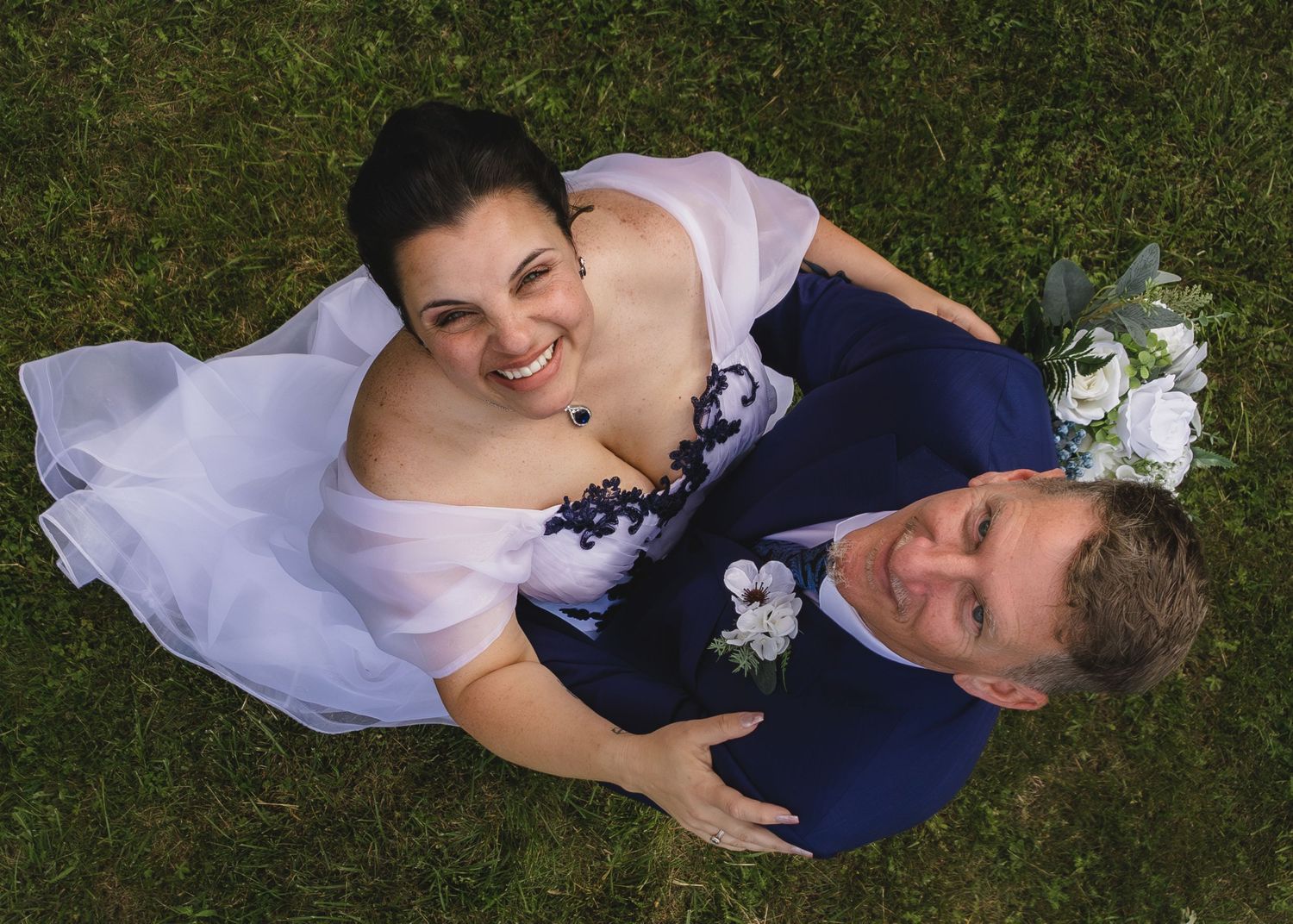 Aerial view of smiling bride in white dress and groom in navy suit lying on green grass holding white bouquet.