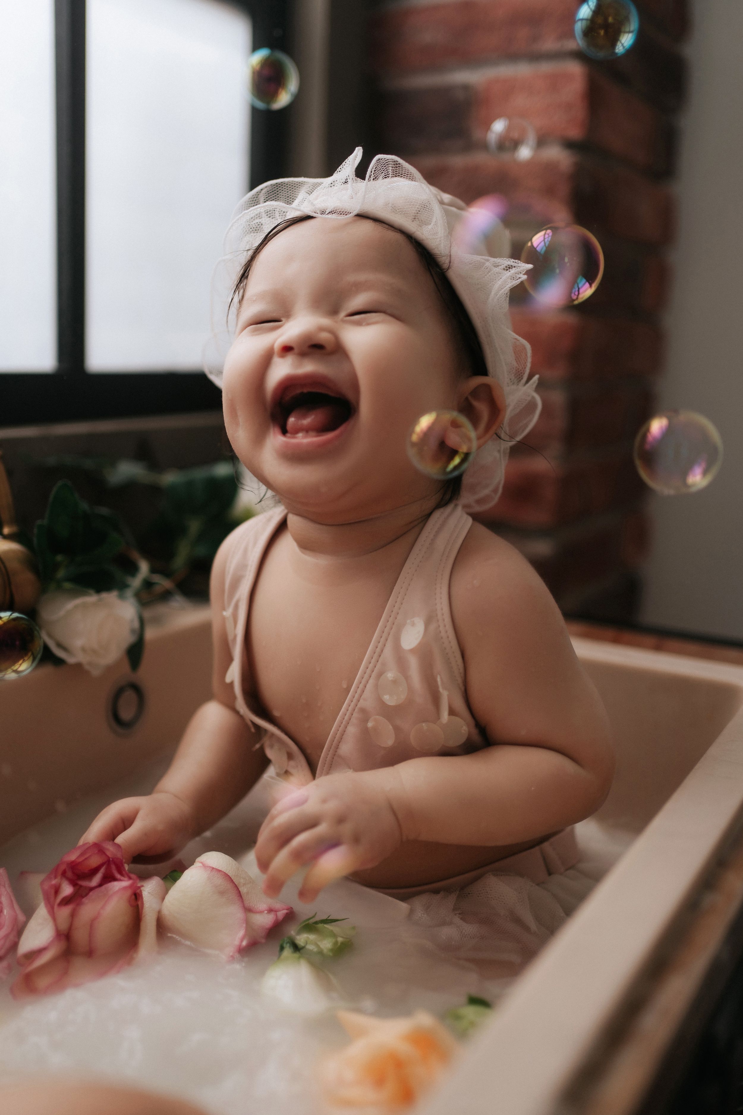 A joyful baby girl laughs in a floral milk bath, wearing a pink outfit and white headband, surrounded by soap bubbles.