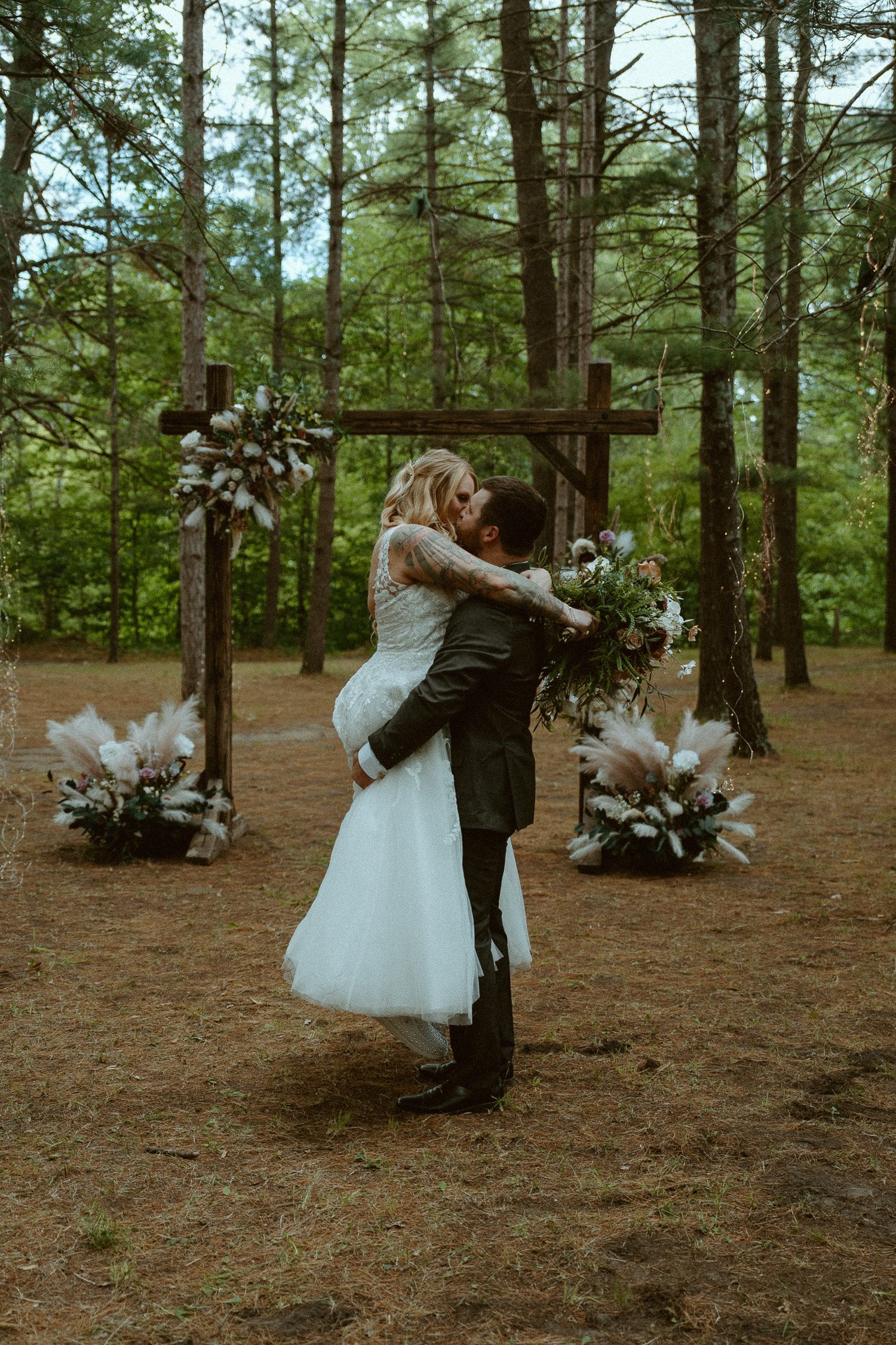 Bride and groom share first kiss in rustic forest wedding ceremony with wooden arch and pampas grass floral arrangements.