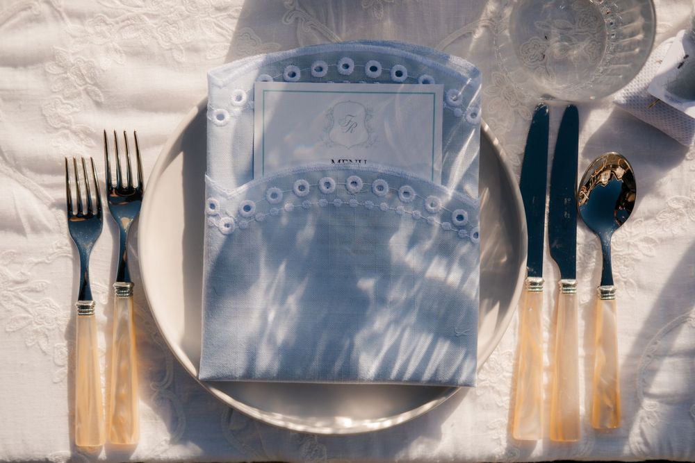 Elegant place setting with blue menu card, silver plate, and gold-handled cutlery on white linen tablecloth.