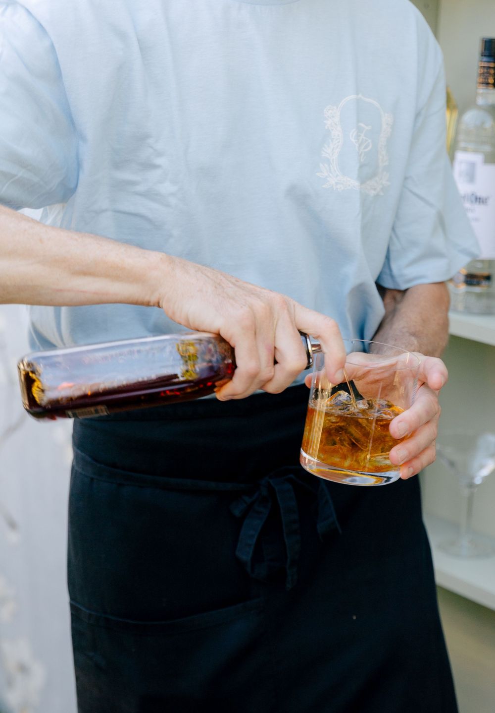 Bartender in blue shirt and black apron pouring whiskey into a rocks glass with ice.
