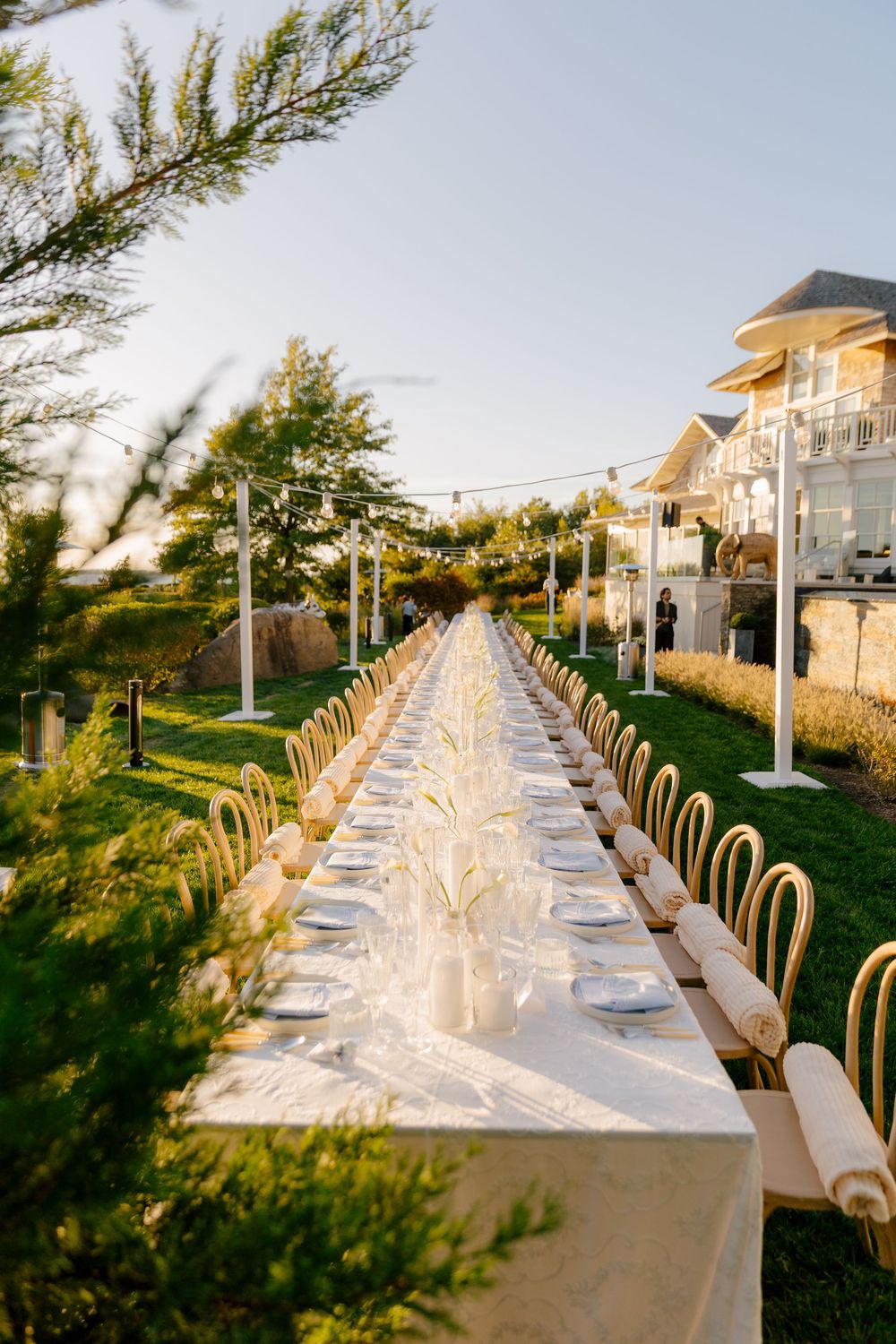 Elegant outdoor long dining table set with white linens and glassware on a green lawn at golden hour.