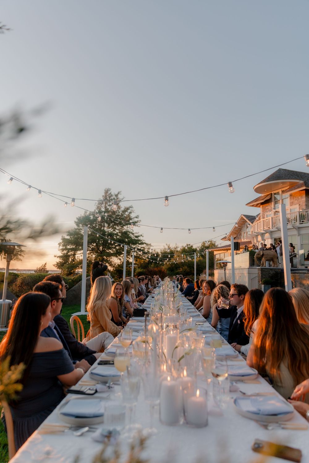 Guests seated at a candlelit long outdoor dining table at sunset with string lights overhead.