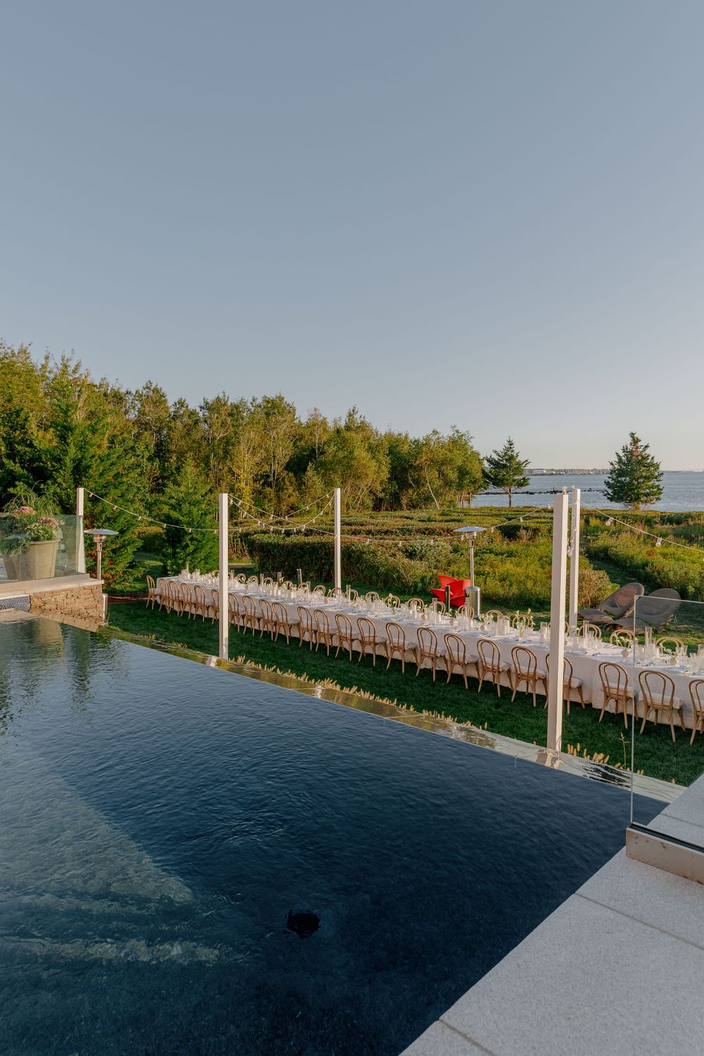Elegant outdoor wedding reception table set alongside an infinity pool with lush greenery and waterfront views at golden hour.