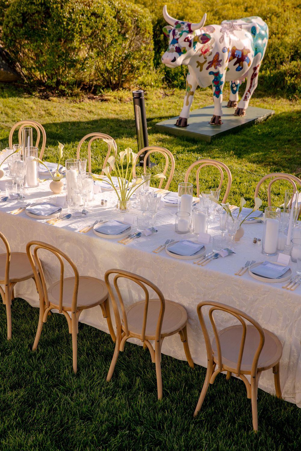 Elegant outdoor dining table set on green grass with gold bentwood chairs and a colorful cow sculpture in background.