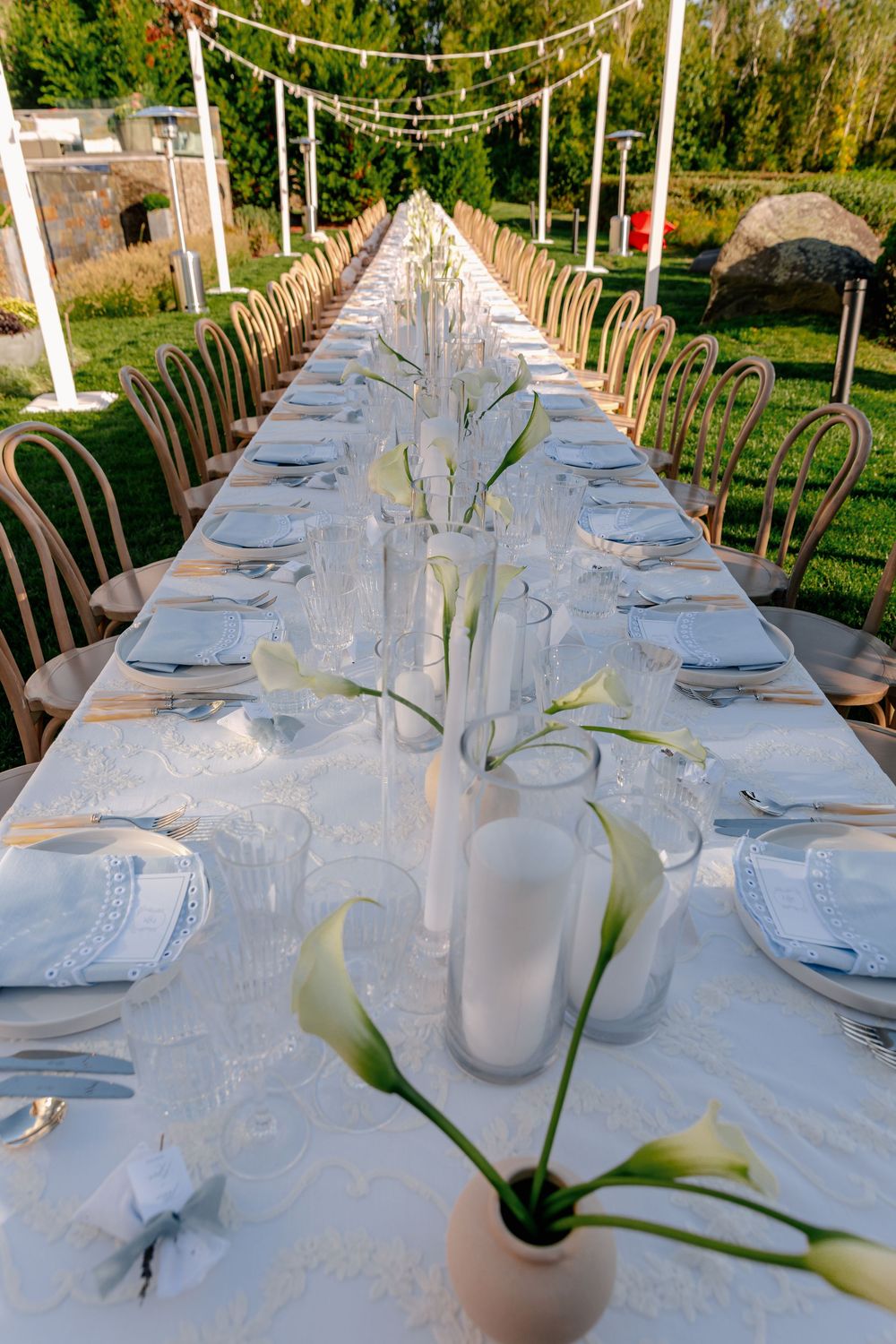 Elegant outdoor wedding reception table set with white linens, calla lilies, candles, and fine dinnerware in a garden setting.