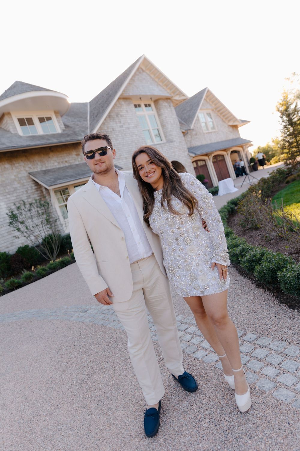 Elegant couple posing in cream outfits before a stone mansion on a paved driveway at dusk.
