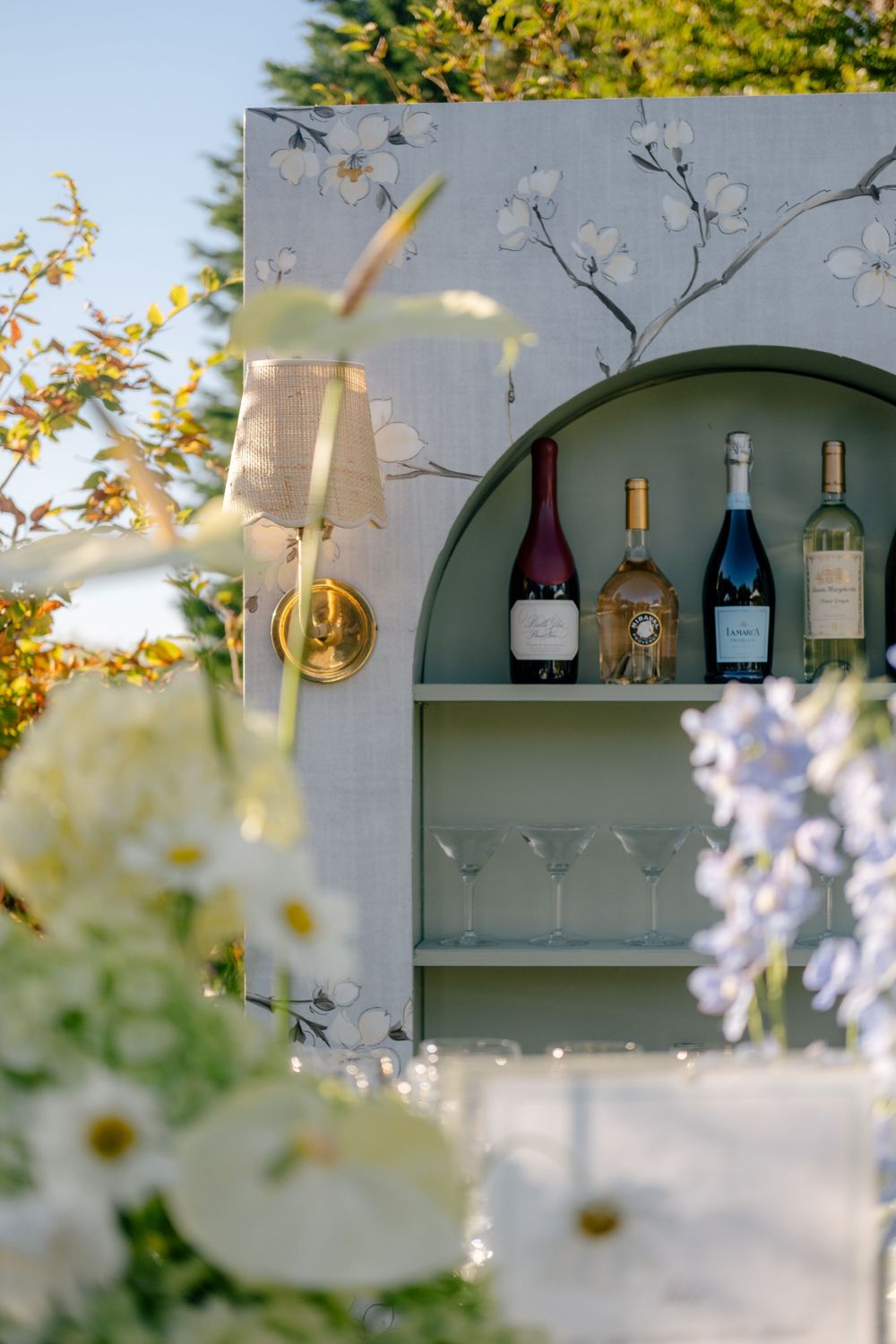 Elegant outdoor bar with floral wallpaper, arched shelf displaying wine bottles and martini glasses, gold wall sconce, and white flowers.