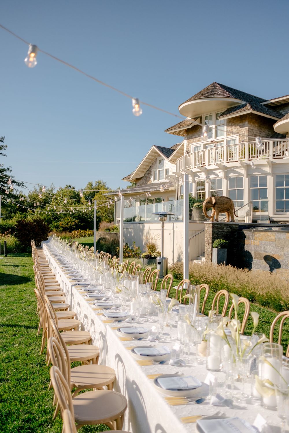 Elegant outdoor wedding reception with long white table, chiavari chairs, string lights, and a grand estate backdrop.
