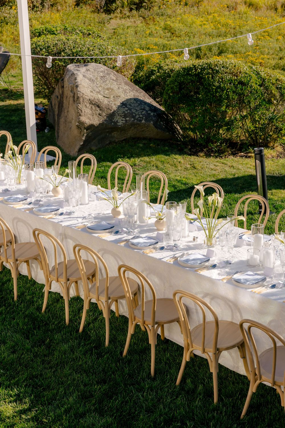 Elegant outdoor wedding reception with long white linen table, bentwood chairs, candles, and floral centerpieces on green lawn.