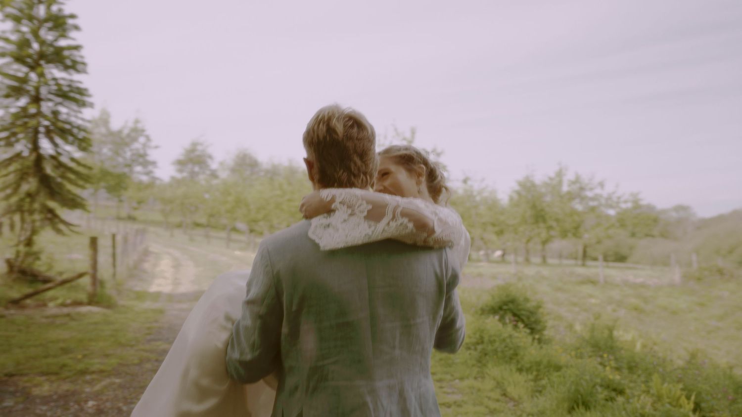 A couple embraces outdoors on a lush green countryside path, the woman in a wedding veil.