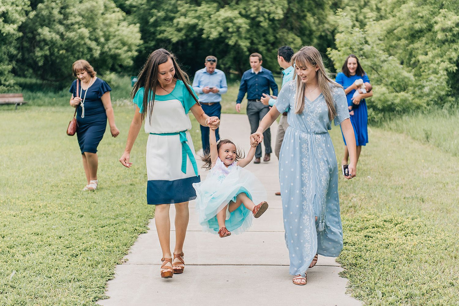 Adults swing a small child while walking together on a paved path through a green summer field.
