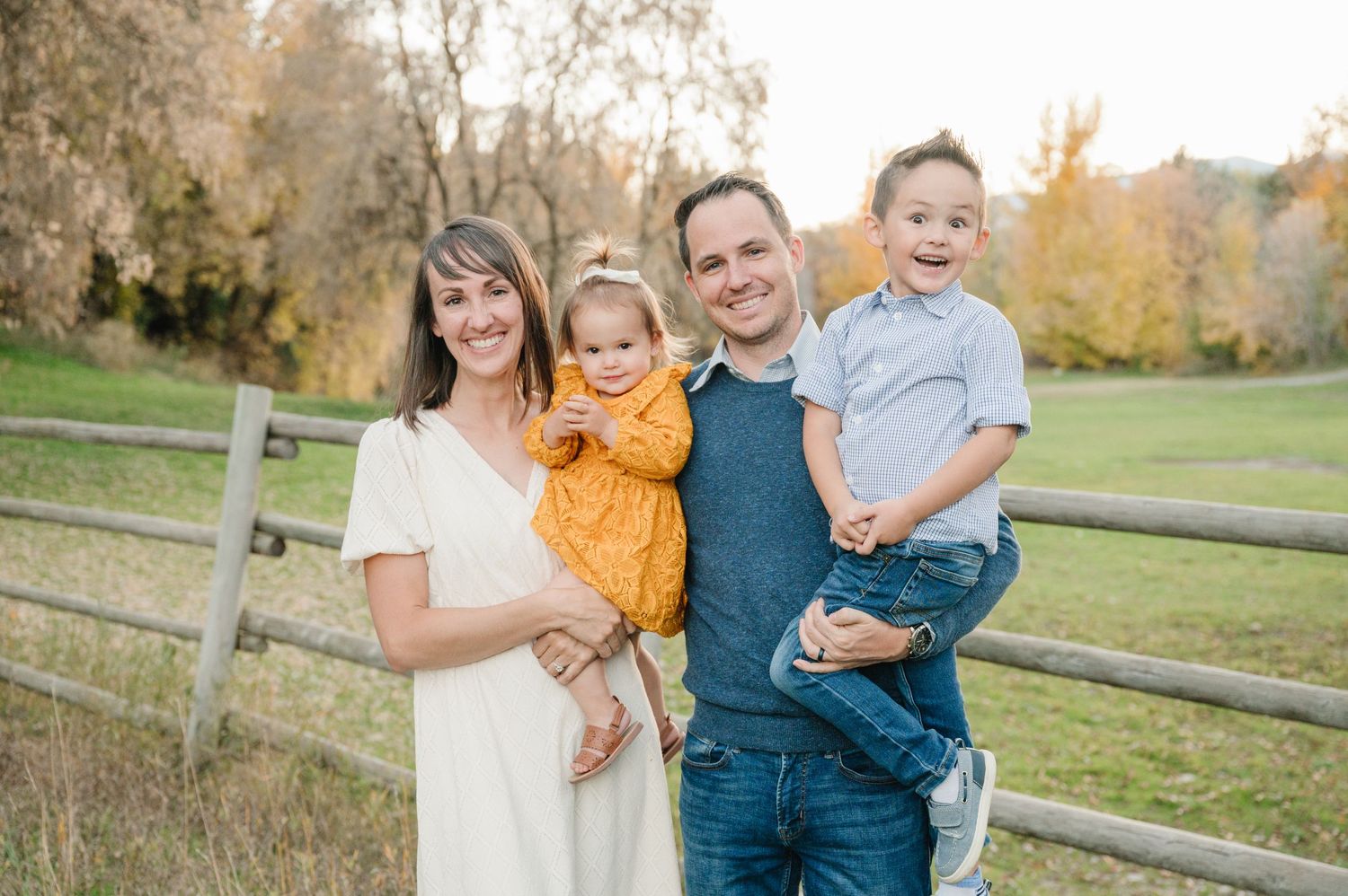 Family of four poses together near a rustic fence during autumn.