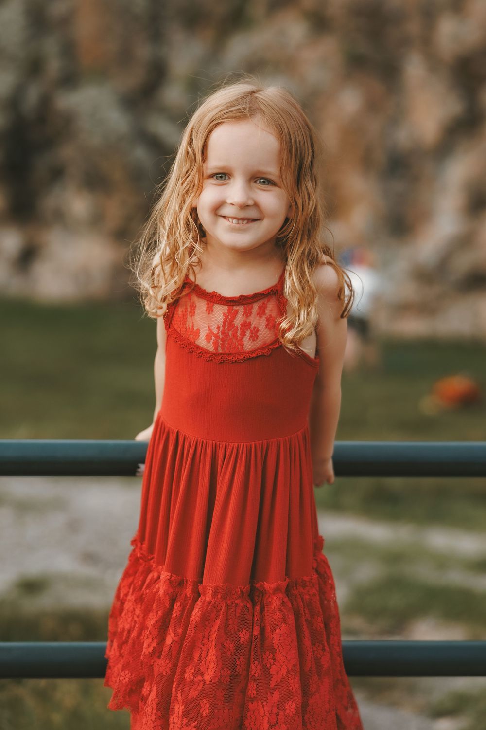 Smiling young girl with curly blonde hair wearing a red lace dress, standing by a railing outdoors.