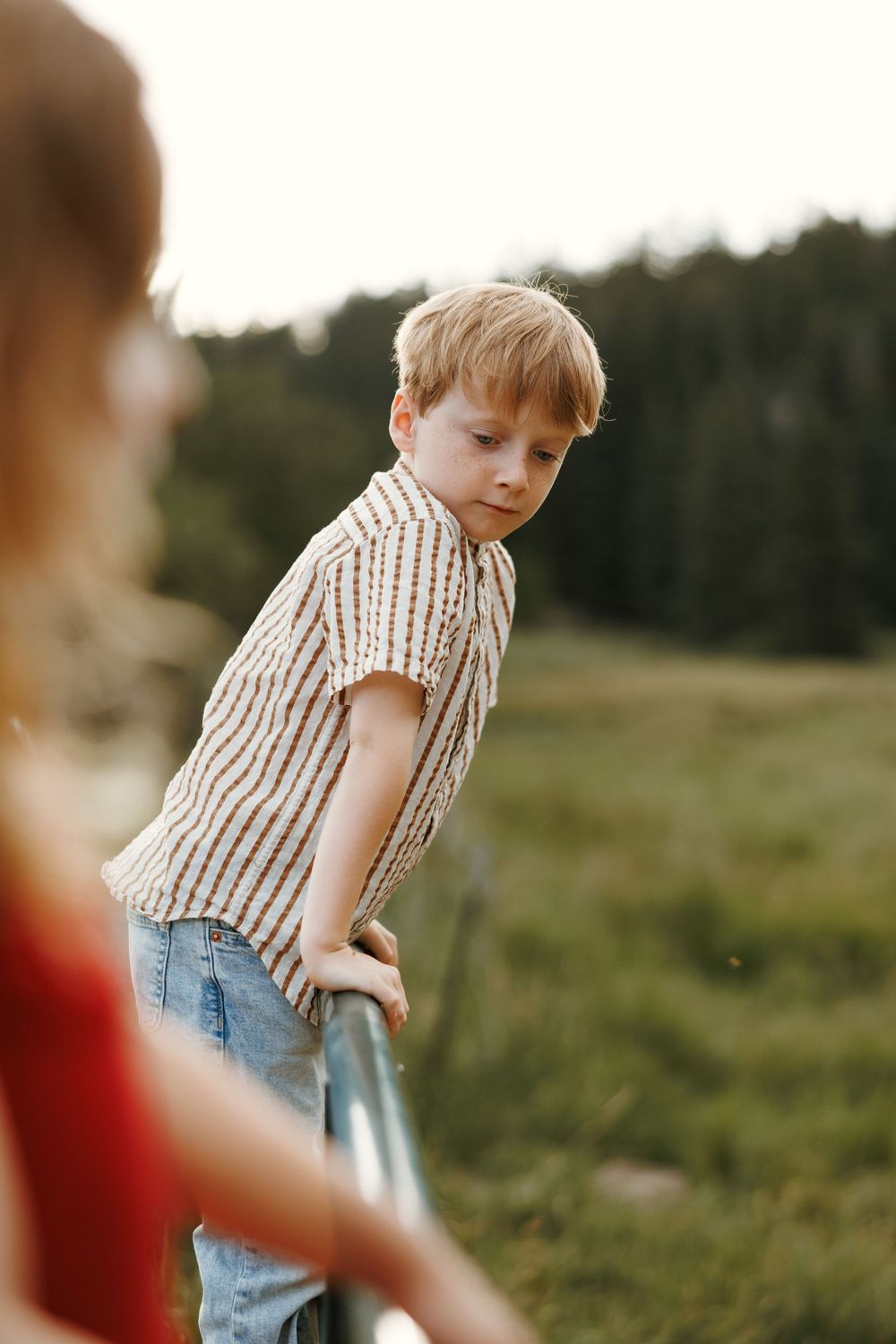 A young blonde boy in a striped shirt leans on a metal railing in a green outdoor field.
