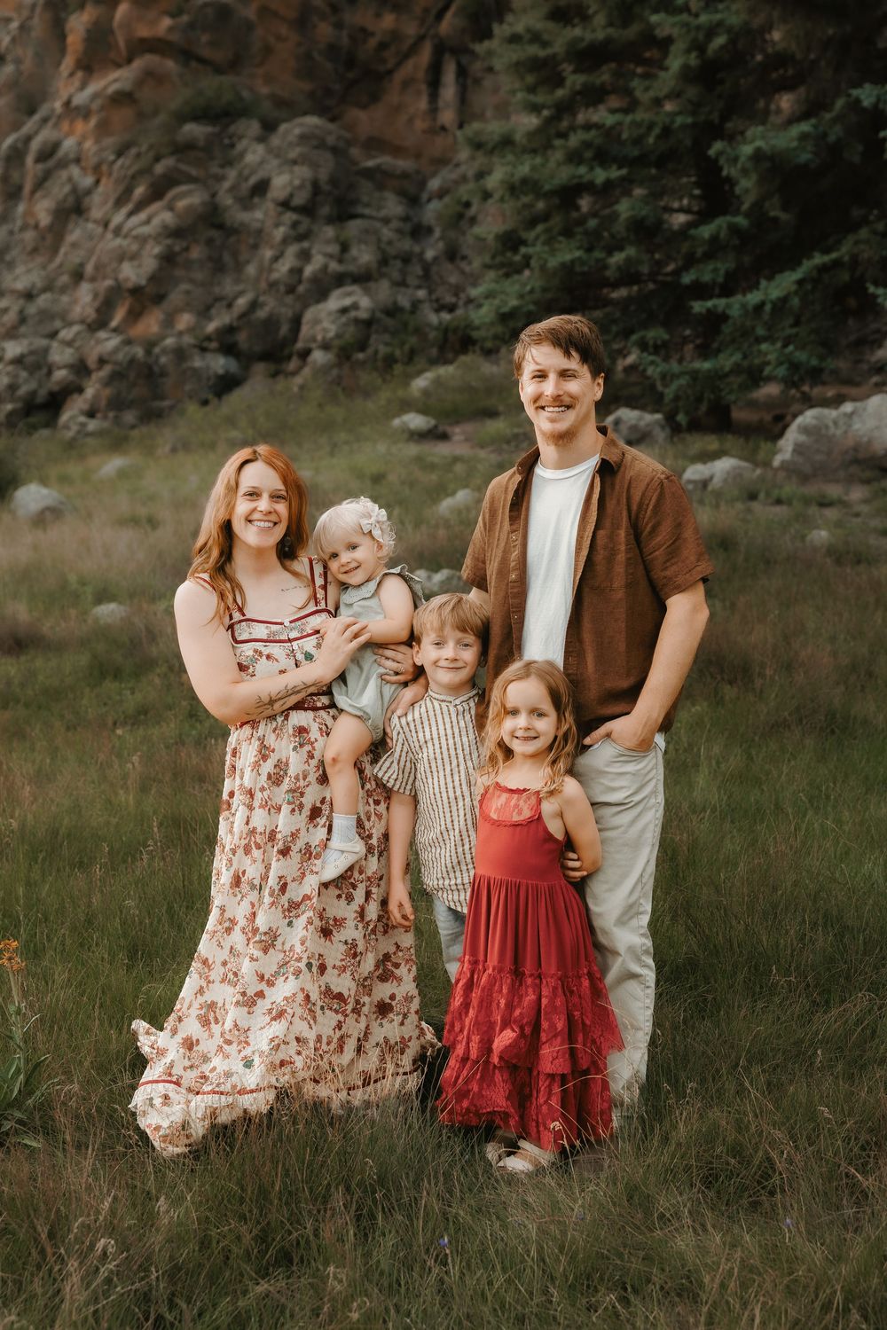Happy family of five posing in a green meadow with rocky cliffs, wearing coordinated red and brown outfits.
