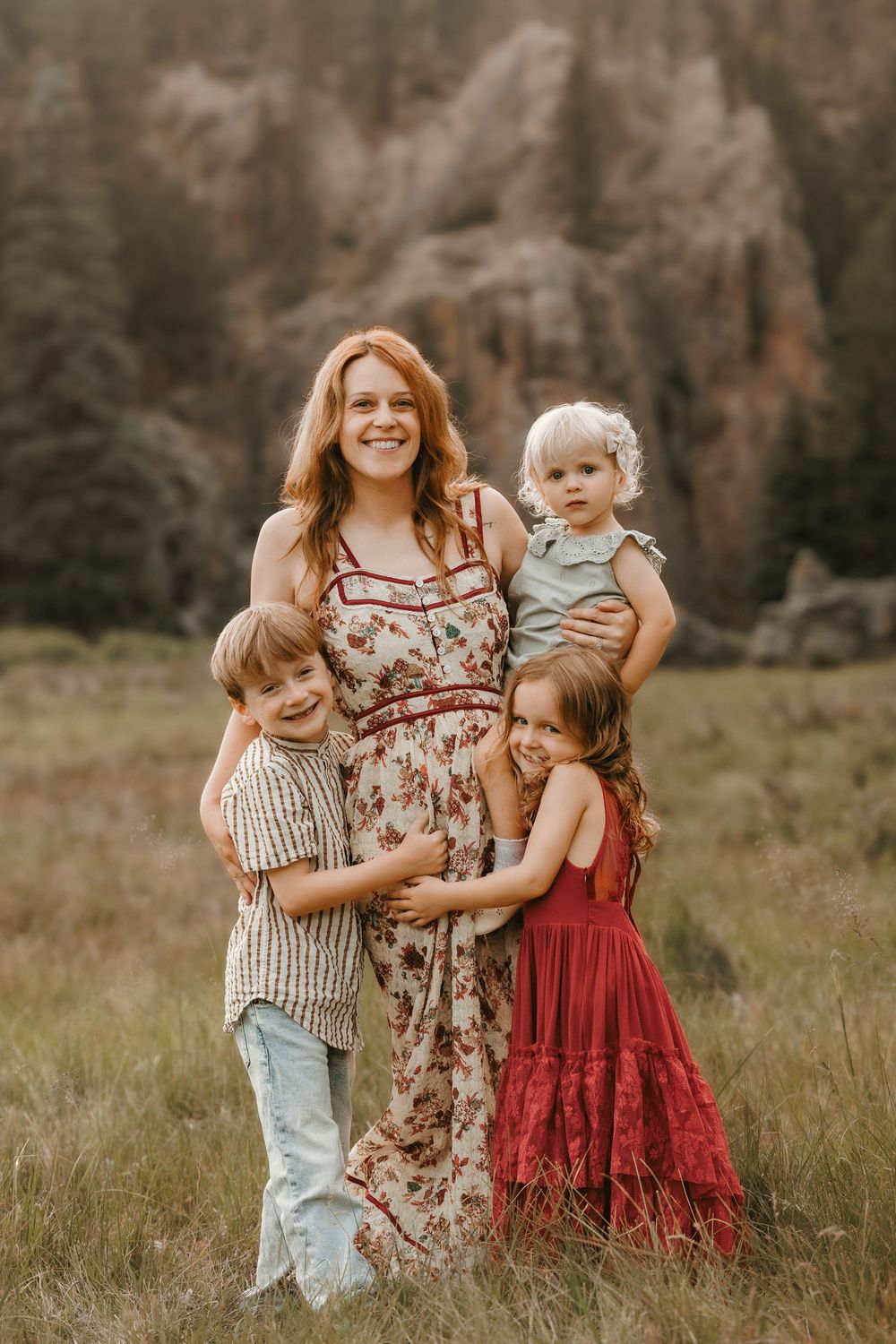A smiling redhead mother in a floral dress poses with her three young children in a mountain meadow.
