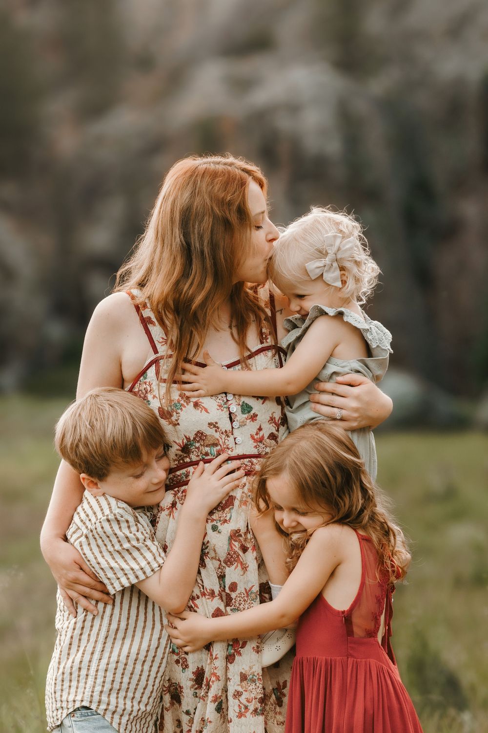 A redheaded mother in a floral dress kisses her toddler while two children embrace her in a scenic outdoor setting.