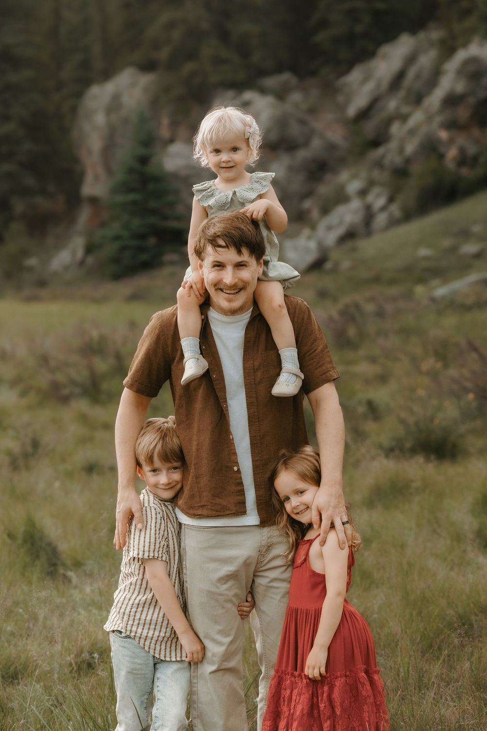 A father smiles outdoors with three young children, one sitting on his shoulders, in a green meadow with rocky cliffs.