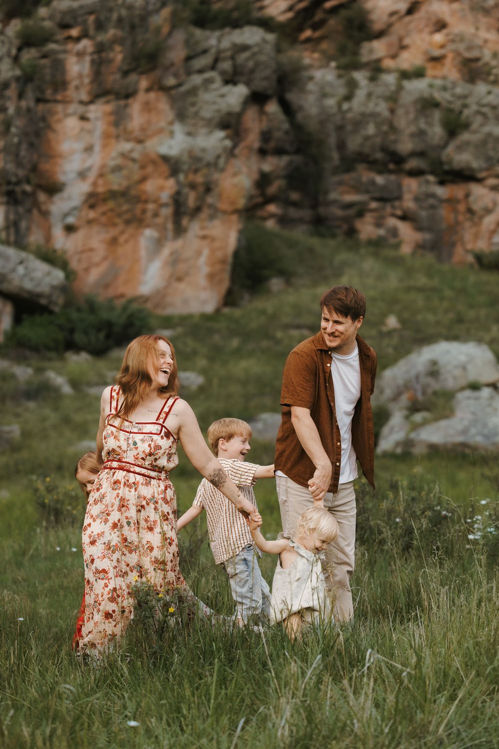 A joyful family plays together in a green meadow with rocky cliffs, mom in floral dress, dad in brown shirt.