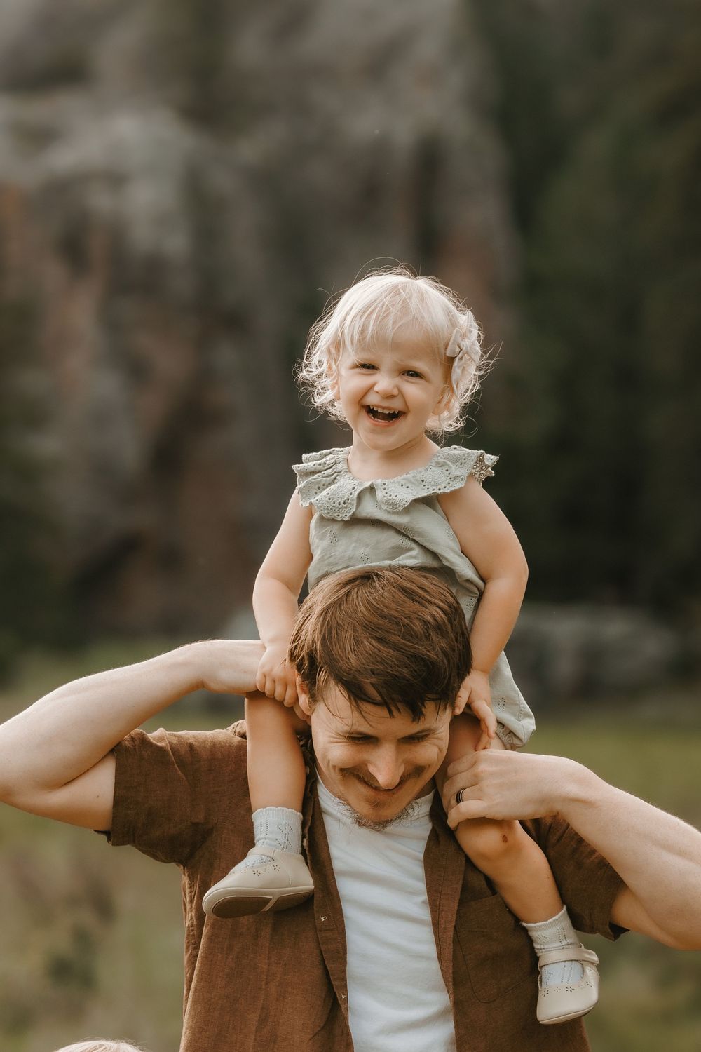 A laughing toddler girl in a green dress sits on a smiling mans shoulders outdoors in a natural setting.