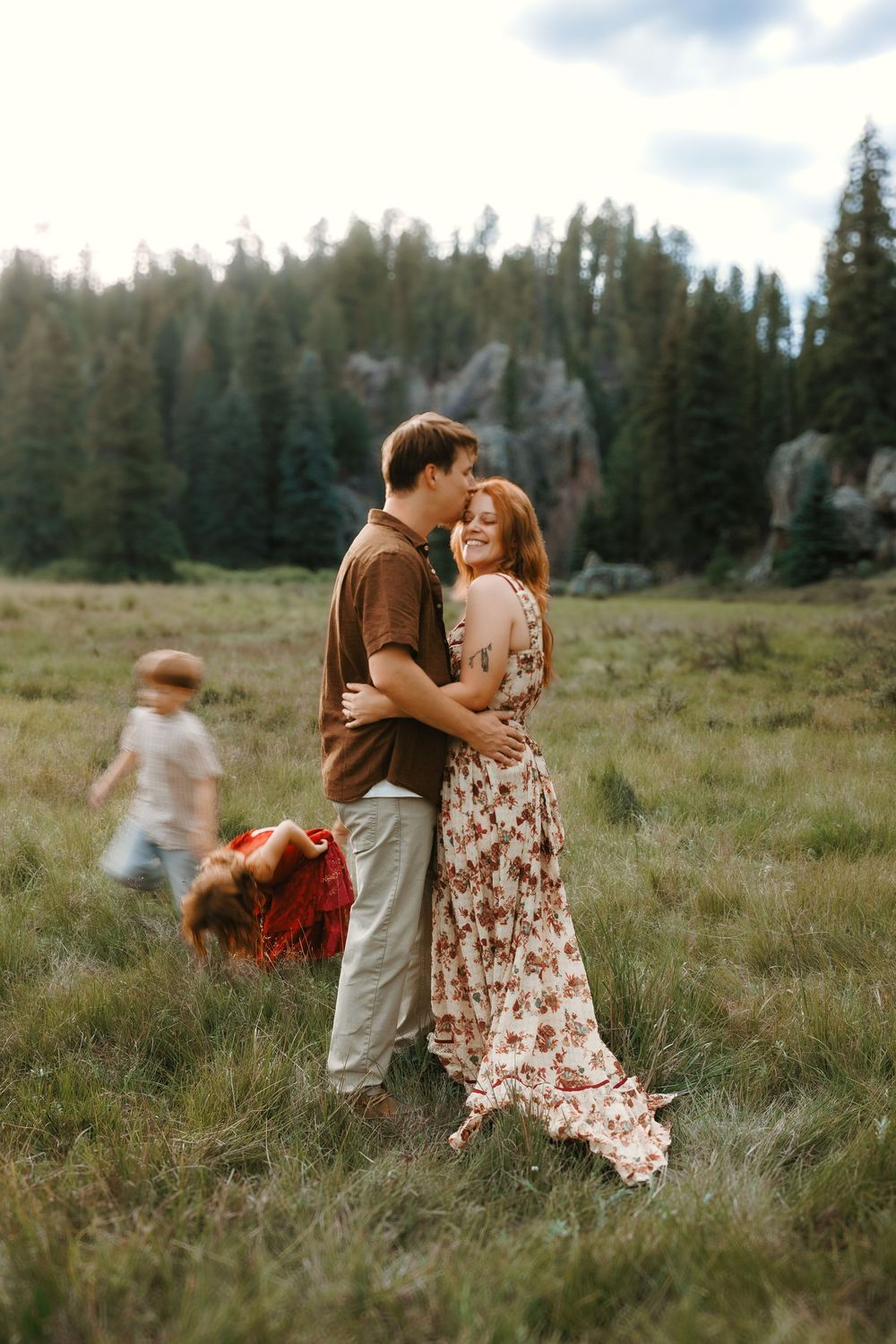 A couple embraces and shares a kiss in a green meadow with pine trees, as children play behind them.