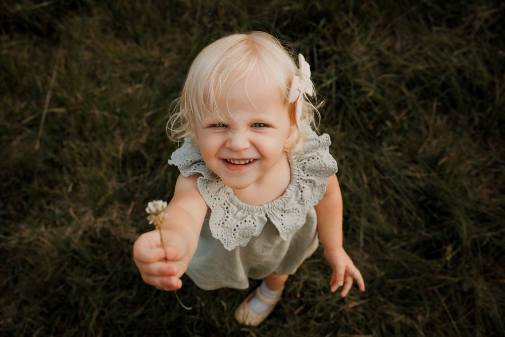 Smiling toddler girl with blonde hair holding a dandelion flower while sitting in green grass outdoors.