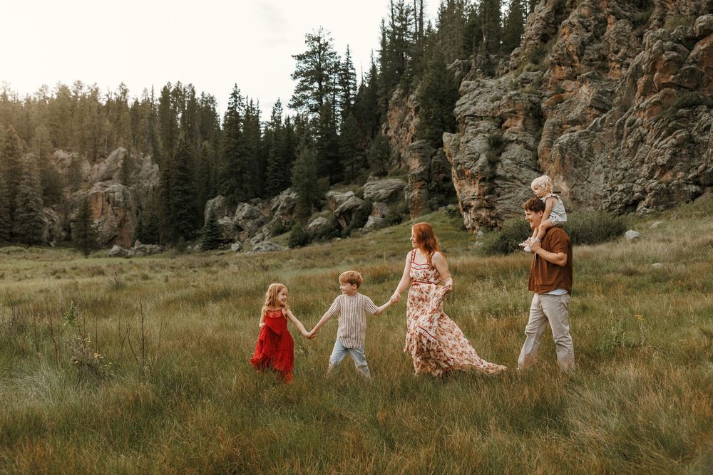 Family of five walking hand-in-hand through a meadow with rocky cliffs and pine trees in golden hour light.